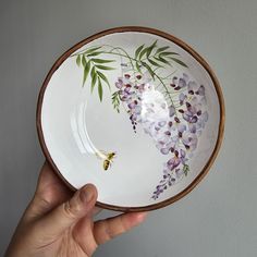 a hand holding a white bowl with purple flowers on the bottom and green leaves painted on it