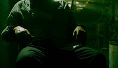 a man sitting in a chair with his hands on his knees, wearing a black shirt and green pants