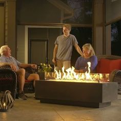 three people sitting around an outdoor fire pit with flames coming out of it and the man standing behind them