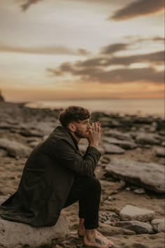 a man sitting on top of a rock next to the ocean holding his hands together