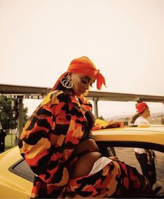 a woman sitting on the hood of a yellow car wearing an orange bandana and earrings