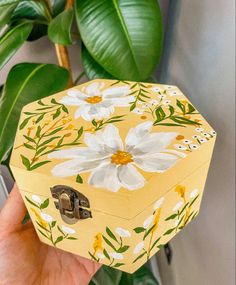 a hand holding a yellow box with white flowers on it and green leaves in the background