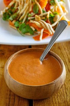 a wooden bowl filled with sauce next to a salad on a white plate and silver spoon