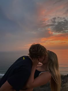 a man and woman kissing while the sun sets over the ocean in front of them