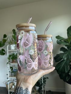 two glass jars with straws in them and butterflies painted on the lids are held up by a person's hand