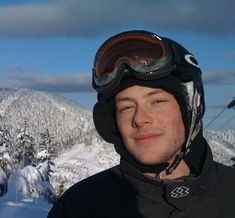 a man with ski goggles on standing in front of snow covered mountains and trees