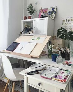 a white desk with lots of art supplies on it and plants in the back ground