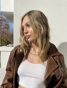 a woman with blonde hair wearing a brown leather jacket and white tank top standing in front of a building