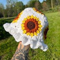 a hand holding up a crocheted sunflower hat in the grass with trees in the background