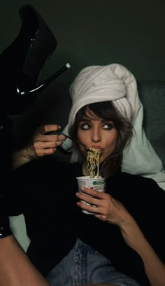 a woman sitting on a couch holding a cup with noodles sticking out of it's mouth