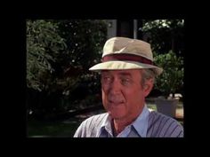 an older man wearing a hat in the middle of a yard with trees and bushes behind him