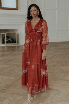 a woman standing in a room wearing a red dress with white flowers and sheer sleeves