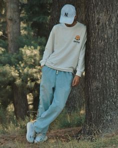 a man leaning against a tree in the woods wearing blue sweatpants and a white hat