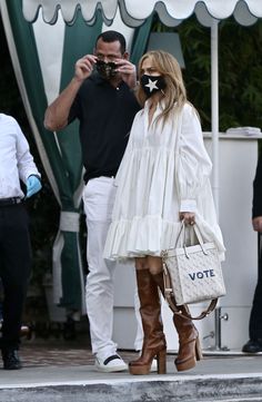 a woman in white dress and cowboy boots taking a photo with her cell phone while another man looks on