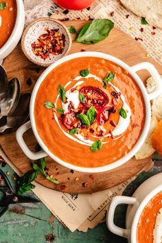 two bowls of tomato soup on a wooden cutting board with bread and tomatoes in the background