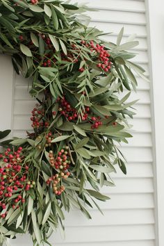 an olive wreath is hanging on the side of a white house with red berries and green leaves