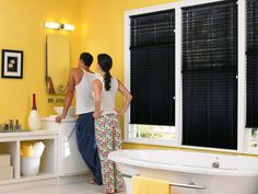 two people standing in front of a bathtub next to a window with black blinds