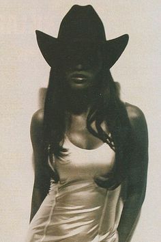 a woman with long hair wearing a cowboy hat and white dress is standing in front of a wall