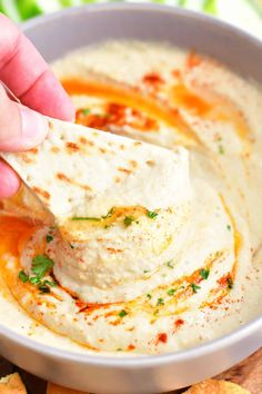 a hand dipping hummus into a white bowl filled with melted cheese and parsley