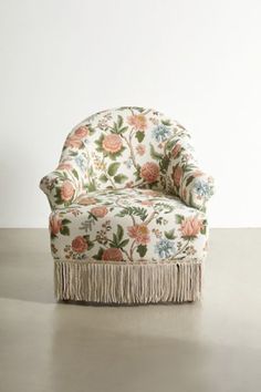 an upholstered chair with fringes on the legs and floral fabric, against a white wall