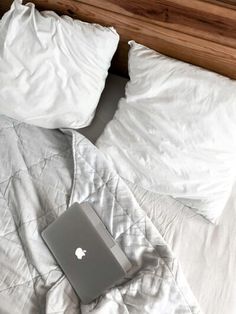 an apple laptop sitting on top of a bed with white comforters and pillows next to it