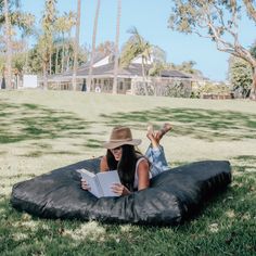 a woman laying on top of a black bean bag chair in the grass reading a book