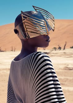 an african woman in a striped shirt and gold headdress stands in the desert