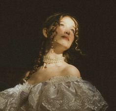 a woman with curly hair wearing a white dress and pearls on her head, looking up