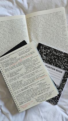 an open book sitting on top of a bed next to a black and white notebook