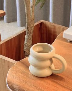 a white coffee cup sitting on top of a wooden table next to a tree in front of a window