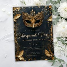 a masquerade party card with white flowers and greenery on the table next to it