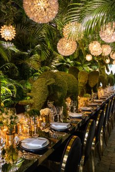 a long table with many plates and place settings on it, surrounded by greenery