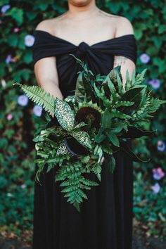 a woman in a black dress holding a bouquet of flowers and greenery on her arm