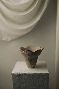 a white vase sitting on top of a block of cement next to a window curtain