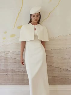 a woman standing in front of a wall wearing a white dress and hat