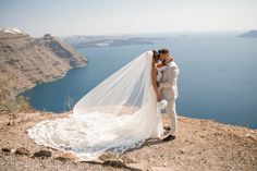 a bride and groom kissing on top of a mountain overlooking the ocean with their veil blowing in the wind