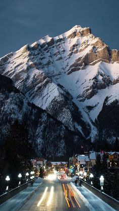 the mountains are covered in snow as cars drive down the road at night with lights on