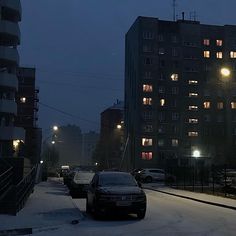 cars parked on the side of a snowy road at night with buildings in the background