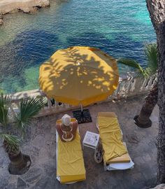 two yellow lounge chairs and an umbrella on the beach next to some water with palm trees