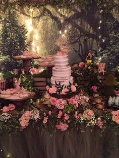 a table topped with a cake surrounded by flowers and fairy land decorations in the background