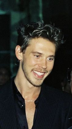 a smiling man in a black shirt and dark suit with his hair pulled back to the side