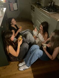 four young women sitting on the floor in a kitchen eating food and drinking beverages together