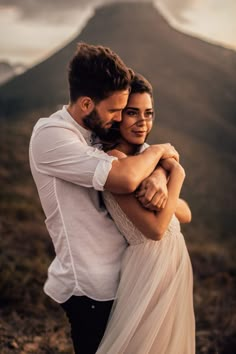 a man and woman hugging each other on top of a hill with mountains in the background