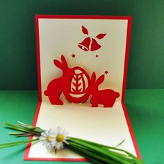 a red and white card with two rabbits on it sitting next to a daisy flower