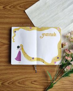 an open book on a wooden table with flowers and a pen in front of it