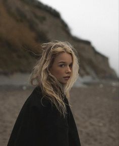 a woman standing on top of a sandy beach next to the ocean with her hair blowing in the wind