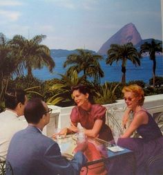 a group of people sitting around a table with water and palm trees in the background