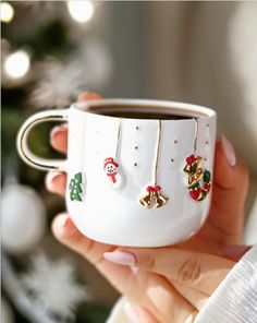 a woman holding a white coffee cup with christmas decorations on the side and gold bells hanging from it