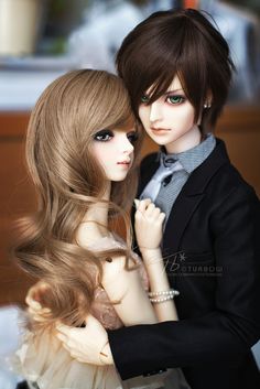 two dolls are posed next to each other wearing formal clothes and hair, one is holding the shoulder of another doll