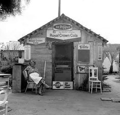 black and white photograph of a man sitting in front of a small building with signs on it